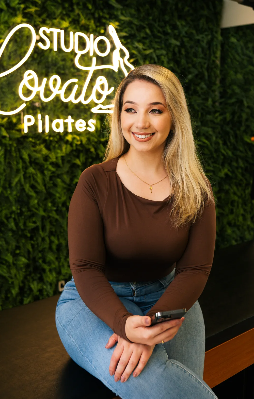 Pricila Lovato sentada no studio, de blusa marrom e jeans, com o telemóvel na mão e o letreiro de neon do Studio Lovato Pilates ao fundo.